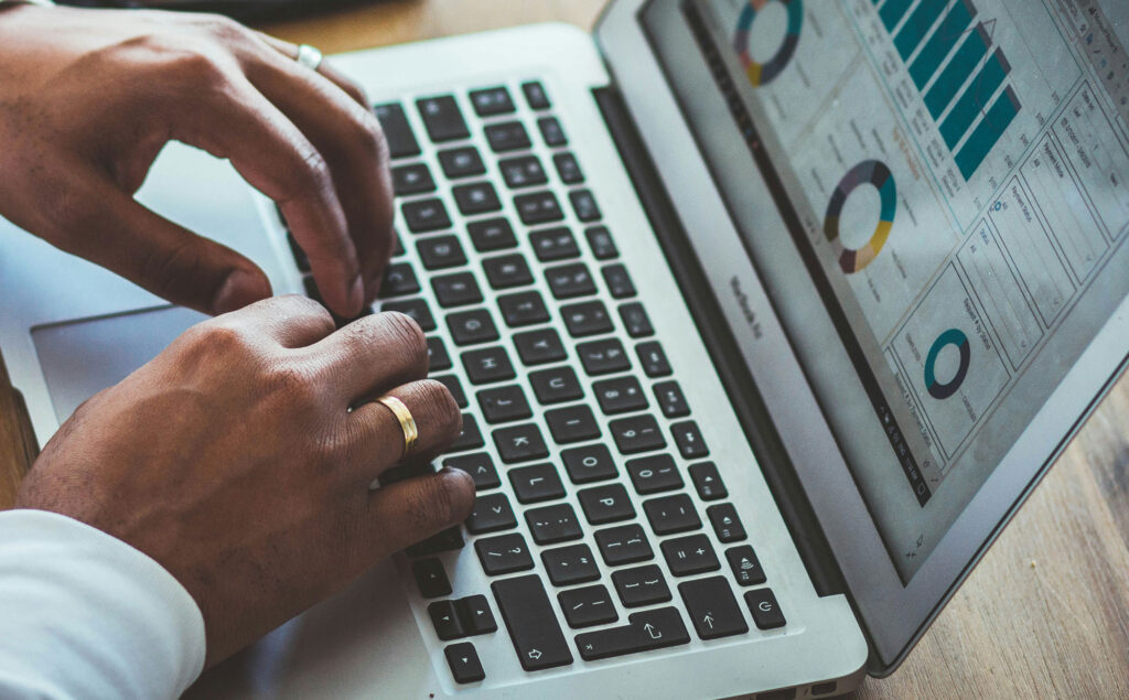 A black man is typing at a laptop. On the screen are various charts.